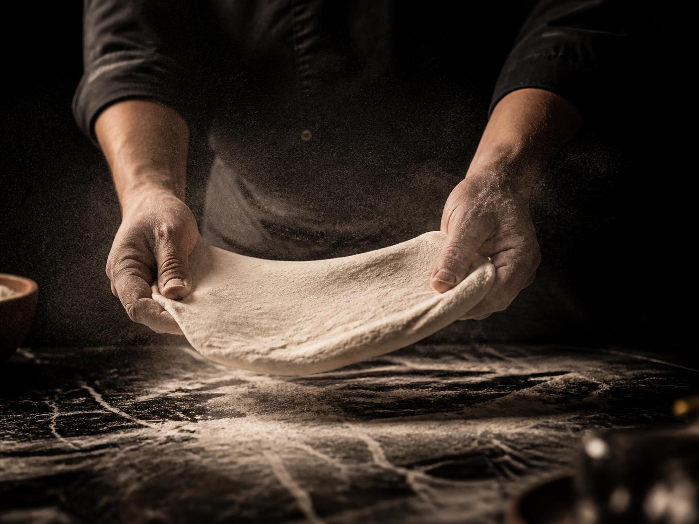 Flour-dusted chef hands stretching pizza dough over a dark marble counter