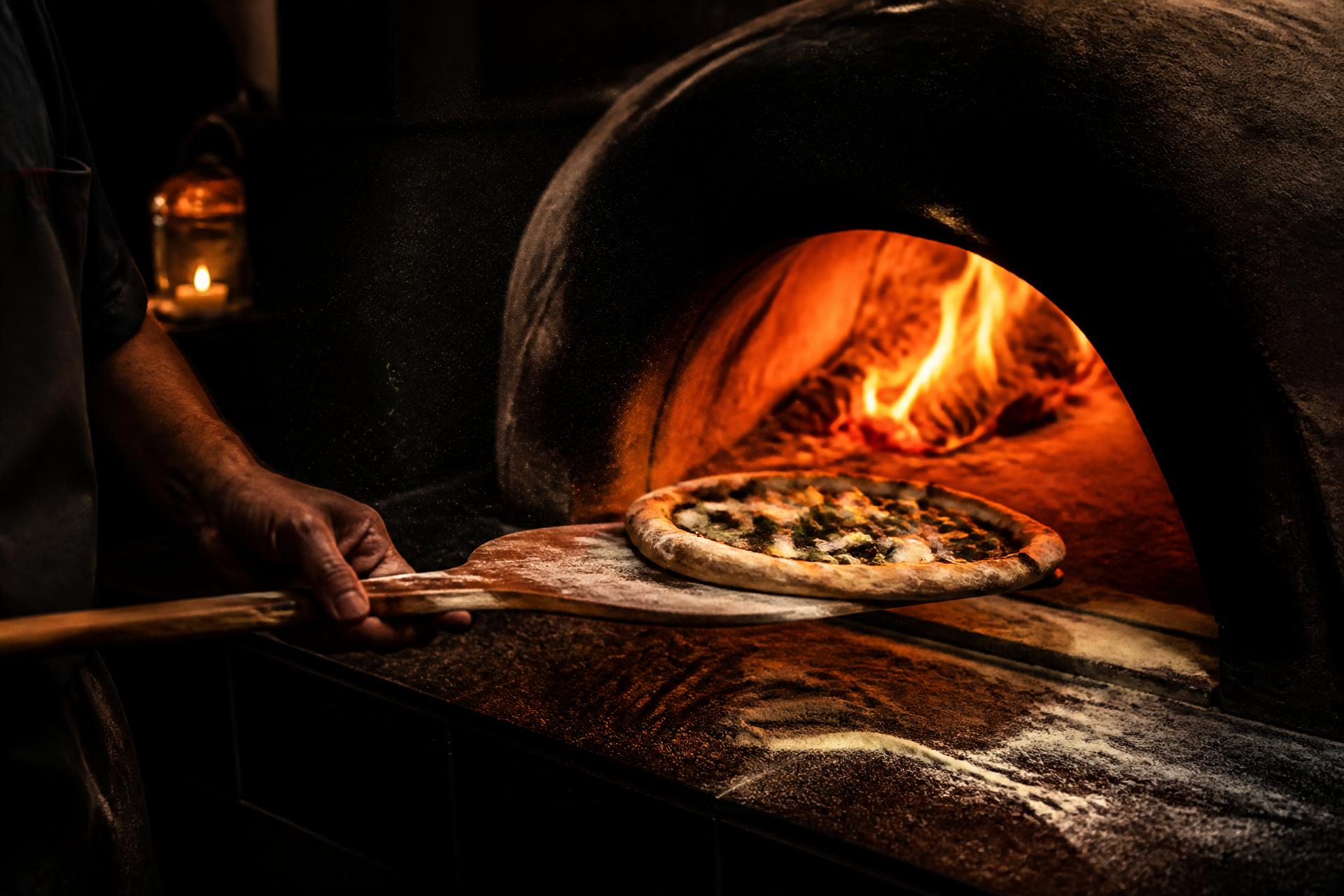 A hand-stretched pizza sliding into a roaring wood-fired oven in a dark, candlelit kitchen