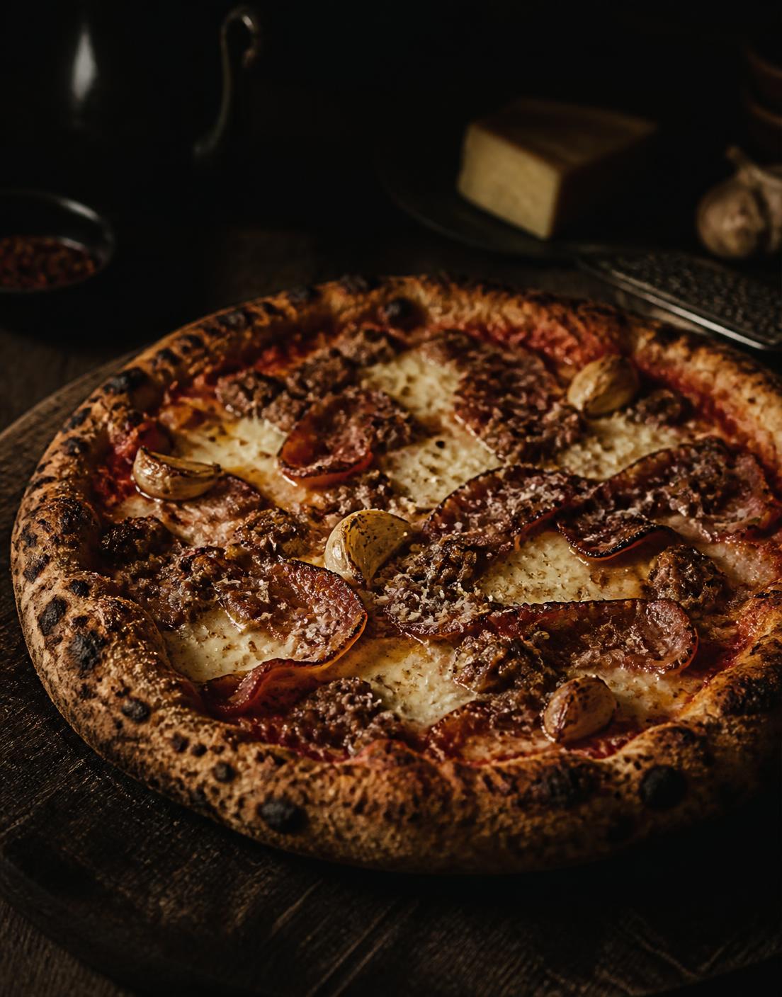 Rustic pizza with fennel sausage, crispy pancetta and roasted garlic on a dark wooden board